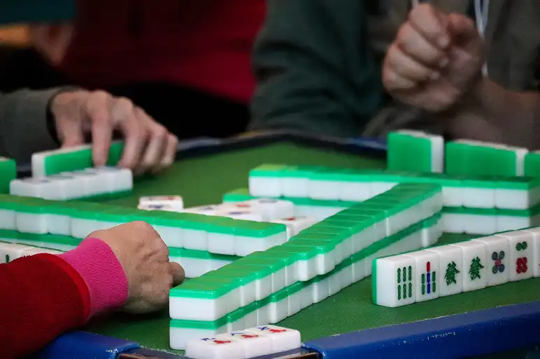 Trzy osoby grające w Mahjong przy zielonym stole, przesuwające płytki rękami. Scena jest skupiona i emanuje koncentracją oraz strategią.
